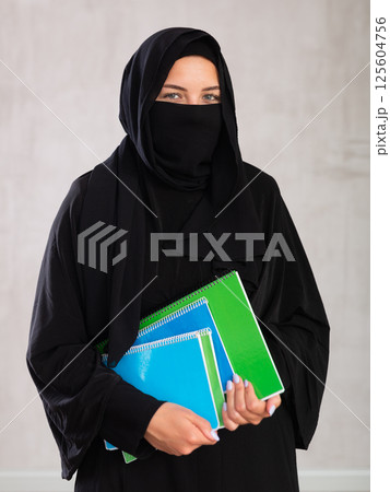 Young student woman dressed in traditional islamic clothes with her face covered holding notebooks Young student woman dressed in traditional islamic clothes with her face covered holding notebooks 125604756