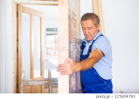 Middle-aged contractor repairman in a blue uniform holding a door in polyethylene before installation in the house 125604765