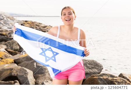 Pretty young girl holding the Israel flag in her hands on the seashore on sunny day 125604771