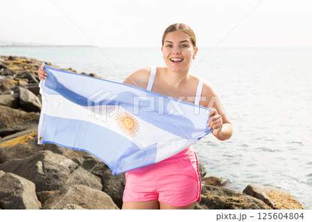 Portrait of happy woman of Argentina match waving a national flag on the seashore on sunny day 125604804