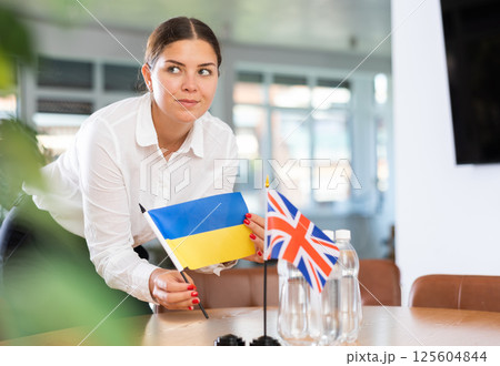 Positive young woman putting little flag of Ukraine on table next to the flag of Great Britain Positive young woman putting little flag of Ukraine on table next to the flag of Great Britain 125604844