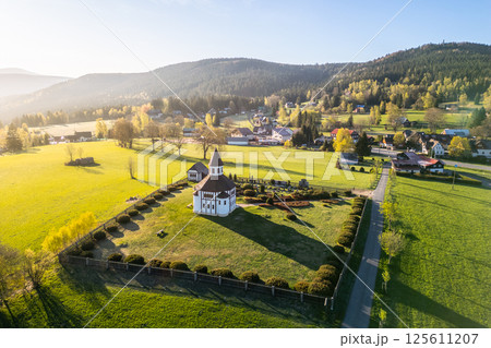 Tesarov Chapel, a striking white octagonal structure, stands amidst lush greenery in Korenov, surrounded by the picturesque Jizera Mountains. The serene landscape basks in soft sunlight. 125611207