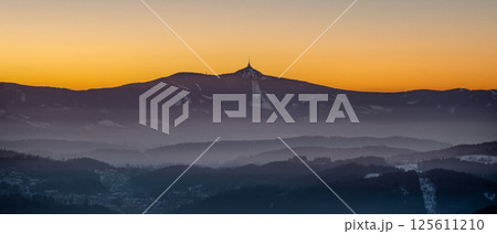 The sun sets behind Jested Ridge, casting a warm orange glow on the silhouette of the landscape. Mist rises from the valleys, creating a serene atmosphere in Czechia's twilight. The sun sets behind Jested Ridge, casting a warm orange glow on the silhouette of the landscape. Mist rises from the valleys, creating a serene atmosphere in Czechia's twilight. 125611210