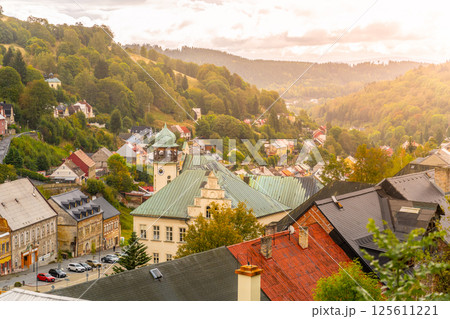 A breathtaking view of Jachymov reveals charming architecture surrounded by lush hills. The sunset bathes the town in warm tones, showcasing its historic buildings and serene atmosphere. 125611221