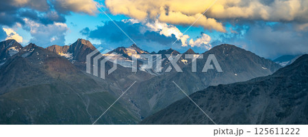 In the evening, the majestic alpine peaks are illuminated by soft light, casting dramatic shadows and highlighting the rugged terrain. Clouds swirl overhead, adding depth to the stunning summer 125611222