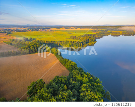 A tranquil morning envelops Rozmberk Pond in Czechia, with soft hazy light filtering through the mist. Surrounding fields and lush greenery reflect in the calm waters, creating a serene atmosphere. 125611235