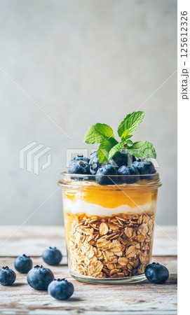Clear glass jar contains layers of granola, yogurt and vibrant yellow fruit puree topped with fresh blueberries and sprig of mint. Healthy and convenient breakfast or snack option 125612326