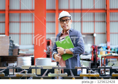Senior Asian industrial engineer is performing inspection a heavy machines and computer systems in the factory. Senior engineer checks manufacturing production line. 125612428