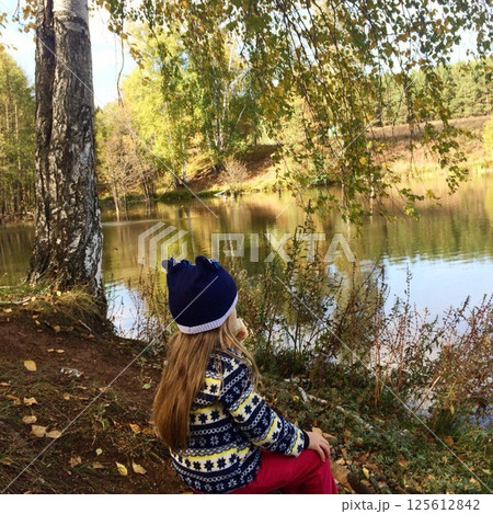 A little girl in a blue hat sits by the river looking at the water. 125612842