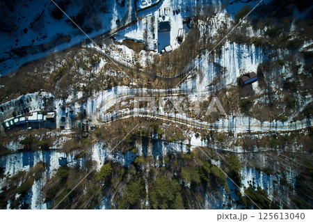 Top-down aerial view of snowy landscape with winding road cutting through forested area. Bare trees cast long shadows on snow, creating intricate patterns. Scattered buildings and vehicles. Top-down aerial view of snowy landscape with winding road cutting through forested area. Bare trees cast long shadows on snow, creating intricate patterns. Scattered buildings and vehicles. 125613040