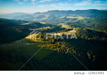 Aerial view of picturesque valley with rolling hills, lush forests, and scattered houses. Sunlight bathes landscape in warm glow, highlighting vibrant greens and creating serene atmosphere. 125613041