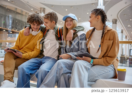 Relaxed teenage friends sitting on bench of big shopping city center Relaxed teenage friends sitting on bench of big shopping city center 125613917