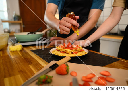 Man drizzling syrup over a stack of homemade pancakes topped with fresh strawberries Man drizzling syrup over a stack of homemade pancakes topped with fresh strawberries 125614226