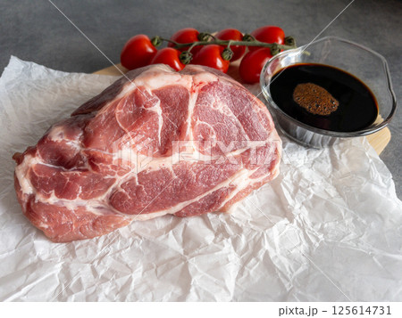 Fresh raw pork meat is placed on crinkled paper alongside ripe cherry tomatoes and cloves of garlic on a rustic wooden board, showcasing an inviting preparation setting. 125614731