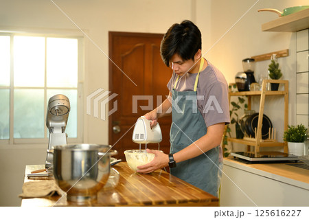 Young asian man using a hand mixer to churn cream in a glass bowl 125616227