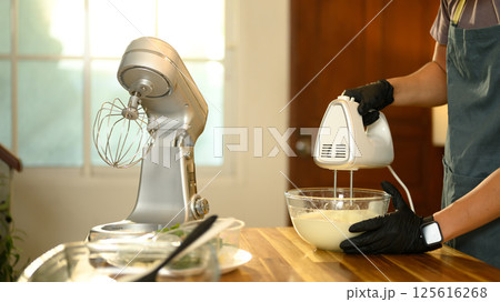 Young asian man using a hand mixer to churn cream in a glass bowl Young asian man using a hand mixer to churn cream in a glass bowl 125616268