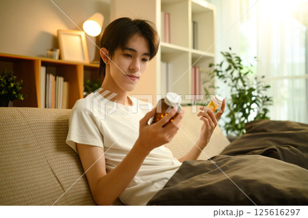 Young man sitting on couch thoughtfully comparing two prescription pill bottles 125616297