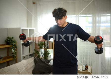Focused man doing shoulder exercises with dumbbells near the sofa 125616309