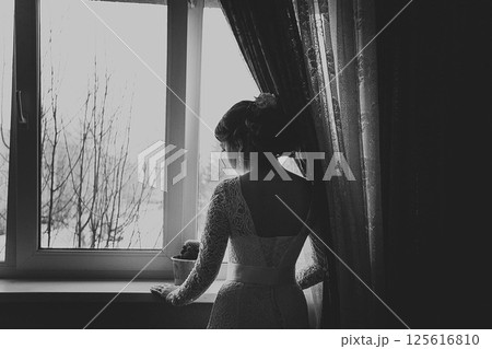 A girl in a wedding dress stands at the window with her back to the viewer. Black and white photo A girl in a wedding dress stands at the window with her back to the viewer. Black and white photo 125616810