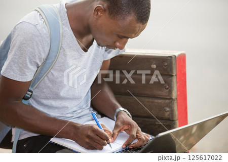 young male student sitting in city working with laptop and notebook 125617022