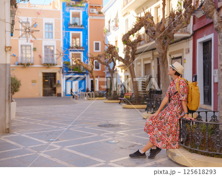 Woman in dress strolls through colorful streets of Spanish coastal town of La Vila Joiosa or Villajoyosa. sunny winter atmosphere highlights charm of Mediterranean architecture and quiet seaside life 125618293