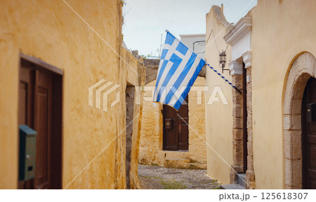 View of Greek flag waving in air serene Mediterranean vibe. beautiful Greek islands series, Narrow cobblestone street lined with charm in downtown Rhodes , Greece. Idyllic stone alley View of Greek flag waving in air serene Mediterranean vibe. beautiful Greek islands series, Narrow cobblestone street lined with charm in downtown Rhodes , Greece. Idyllic stone alley 125618307