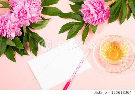 Minimalistic greeting card with pen and tea, set against vibrant peonies and soft pink background. 125618364