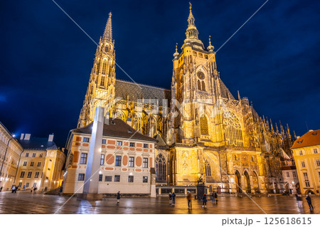 St Vitus Cathedral stands majestically at night, bathed in golden light, surrounded by the historic Prague Castle. Visitors stroll through the serene square, admiring the stunning architecture. 125618615