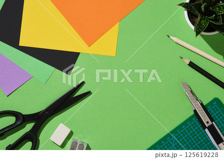Frame of supplies for paper art hobby: scissors, craft knife, pencil, eraser, cutting mat, colored paper on green background. Copyspace. Frame of supplies for paper art hobby: scissors, craft knife, pencil, eraser, cutting mat, colored paper on green background. Copyspace. 125619228