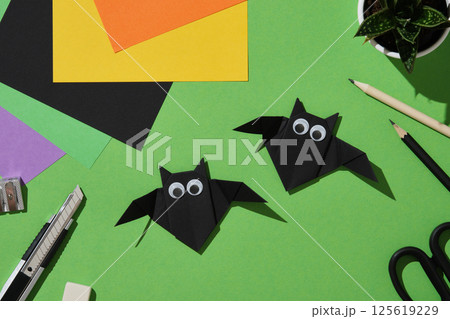 Two black paper handmade origami origami bats with googly eyes are arranged on a green background surrounded by colorful sheets, pencils, scissors, cutter, eraser and sharpener. Two black paper handmade origami origami bats with googly eyes are arranged on a green background surrounded by colorful sheets, pencils, scissors, cutter, eraser and sharpener. 125619229