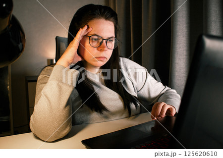 Woman working on laptop late evening at home. Concentrated female employee solves work issues sitting at computer at work desk. Woman working on laptop late evening at home. Concentrated female employee solves work issues sitting at computer at work desk. 125620610