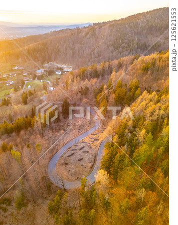 Above view of a winding road through a forest during sunset. Golden hues illuminate the trees and landscape, creating a tranquil, picturesque setting perfect for enjoying nature. 125621553