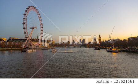 The London Eye stands tall on the South Bank, overlooking the River Thames as boats glide by. The setting sun casts a warm glow, highlighting the iconic skyline of London. 125621561