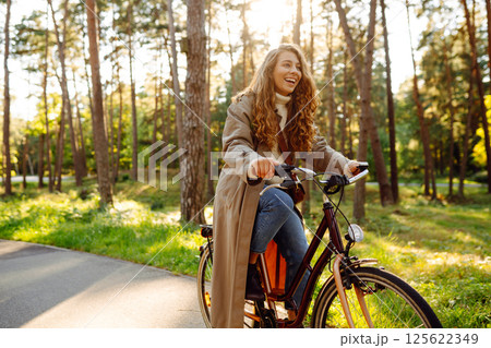 Young happy woman in yellow coat rides bicycle in sunny park. Woman enjoys autumn nature. Lifestyle. 125622349
