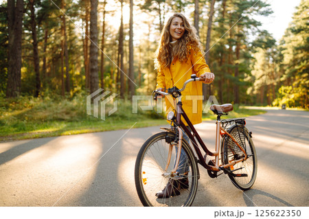 Young happy woman in yellow coat rides bicycle in sunny park. Woman enjoys autumn nature. Lifestyle. 125622350