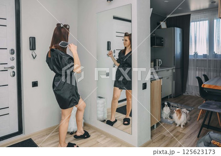 woman brushing hair in front of full-length mirror in black robe with reflection, japanese chin dog on wooden floor in modern home interior, beauty, home living, pet lifestyle 125623173