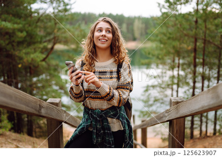 A young woman in a cozy sweater walking near a beautiful lake, smiling and using her phone A young woman in a cozy sweater walking near a beautiful lake, smiling and using her phone 125623907