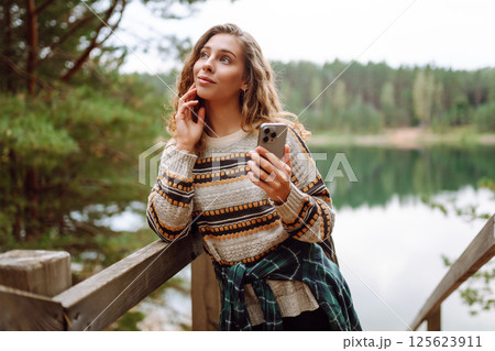 A young woman in a cozy sweater walking near a beautiful lake, smiling and using her phone A young woman in a cozy sweater walking near a beautiful lake, smiling and using her phone 125623911