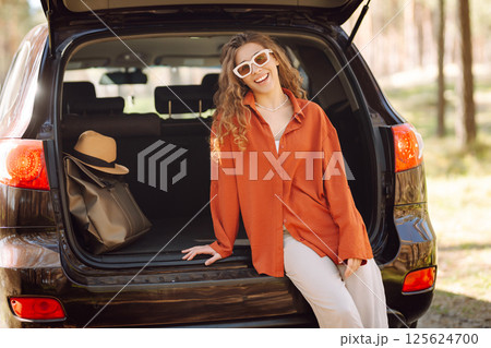 A traveler young woman smiles while sitting in the trunk of her car during a sunny day in a forest A traveler young woman smiles while sitting in the trunk of her car during a sunny day in a forest 125624700