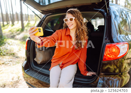 Happy woman sits on the trunk of her car and taking a selfie. Selfie while traveling by car. 125624703