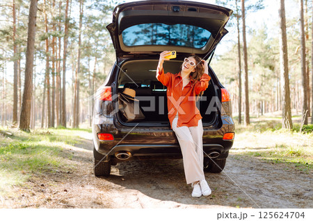 Happy woman sits on the trunk of her car and taking a selfie. Selfie while traveling by car. 125624704
