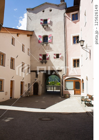 Historic buildings with colorful shutters surrounding a stone-paved courtyard leading to an arched passageway and glimpses of greenery beyond Historic buildings with colorful shutters surrounding a stone-paved courtyard leading to an arched passageway and glimpses of greenery beyond 125626149