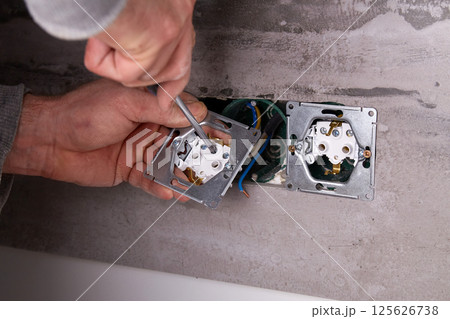 An electrician uses a screwdriver to install an electrical outlet on a wall covered with porcelain stoneware 125626738