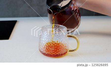 Pouring tea from a glass teapot into a textured glass mug with a yellow handle on a white countertop in a contemporary kitchen, setting a cozy and welcoming ambiance Pouring tea from a glass teapot into a textured glass mug with a yellow handle on a white countertop in a contemporary kitchen, setting a cozy and welcoming ambiance 125626895