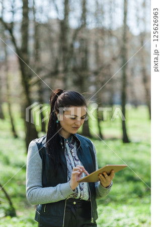 Girl in leather pants and bomber jacket with a tablet in the park. Beautiful girl is smiling and looking at the tablet. 125626963