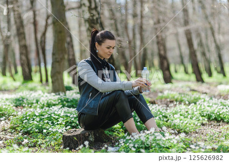Black-haired woman opens a plastic bottle of water in a spring forest sitting on a tree stump. Beautiful girl in the park with a bottle of water sitting on a tree stump. Black-haired woman opens a plastic bottle of water in a spring forest sitting on a tree stump. Beautiful girl in the park with a bottle of water sitting on a tree stump. 125626982