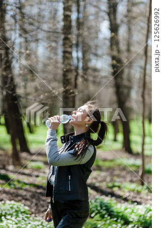 Black-haired woman drinking water from a plastic bottle in the forest. A beautiful girl in a green park drinking water from a bottle. Black-haired woman drinking water from a plastic bottle in the forest. A beautiful girl in a green park drinking water from a bottle. 125626992