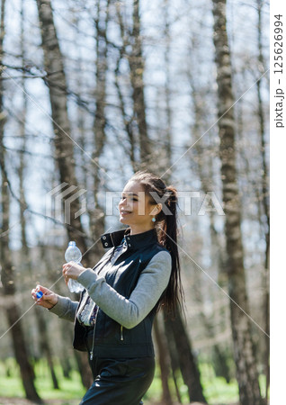 A beautiful girl in leather pants and a bomber jacket holds a bottle of water in a spring forest and basks in the sun. A young woman is walking in the park with a bottle of mineral water. 125626994