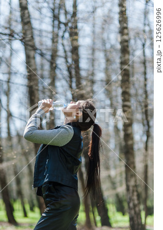 Woman with a long tail drinking water in a green forest. Young girl drinking water in the park.  125626996