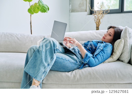 Young serious woman typing on laptop lying on couch at home 125627081